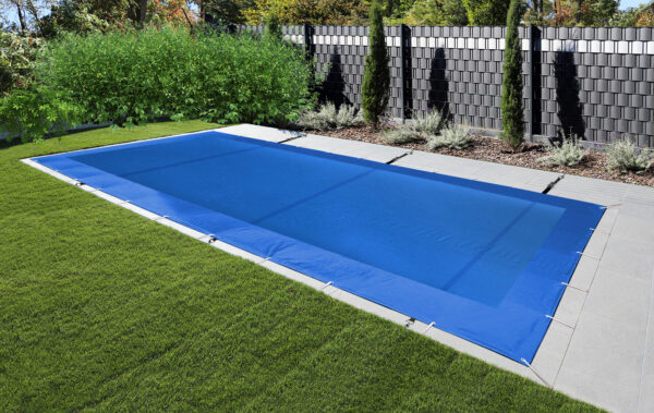 Ein eleganter Außenbereich verfügt über einen rechteckigen Pool mit blauer Abdeckung, umgeben von grünem Gras und einem gepflasterten Bereich, mit einer Reihe hoher Pflanzen und einem modernen dunklen Zaun. Der Stabilisierungsgurt vervollständigt die Einrichtung.