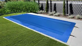 Ein mit einer blauen Plane abgedeckter Pool ist von Gras und grauen Steinfliesen umgeben, im Hintergrund sind hohe Pflanzen und ein moderner schwarzer Stabilisierungsgurtzaun zu sehen.