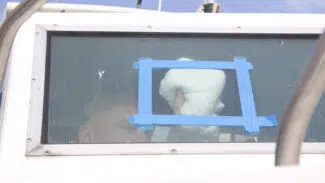 Eine Person verwendet den Vuplex Kunststoffreiniger (Klarsichtfolien-Reiniger), um ein Fenster durch eine mit blauem Klebeband versehene rechteckige Öffnung an der Scheibe eines Bootes oder Fahrzeugs zu reinigen.