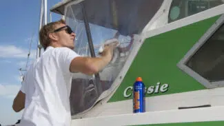 Eine Person mit Sonnenbrille putzt das Fenster eines grün-weißen Bootes namens Cassie mit einem Tuch, während in der Nähe eine Flasche Vuplex Kunststoffreiniger (Klarsichtfolien-Reiniger) steht.