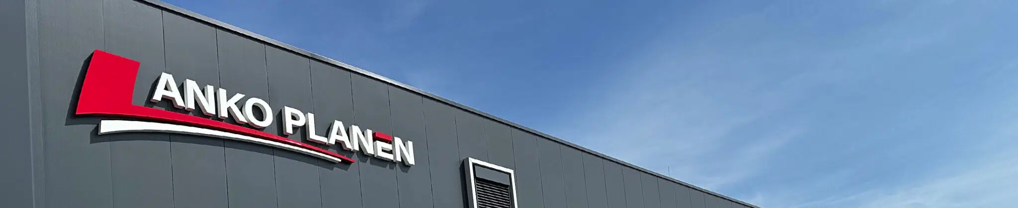 Ein modernes graues Gebäude mit dem ANKO PLANEN-Logo in großen roten und weißen Buchstaben an der Wand, vor einem klaren blauen Himmel.