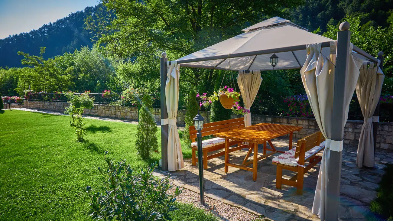 Ein hölzerner Picknicktisch und Bänke stehen unter einem beigen Baldachin mit Vorhängen in einem üppigen Garten. Bäume und ein Holzzaun säumen den Hintergrund und schaffen eine ruhige, sonnige Umgebung.