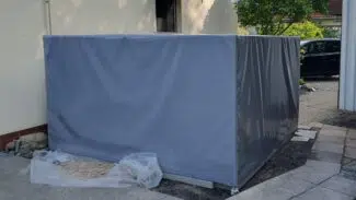 Ein großer rechteckiger Gegenstand, der mit einer rechteckigen Abdeckhaube abgedeckt ist, steht neben einem cremefarbenen Gebäude mit Plastik und Sand in der Nähe.