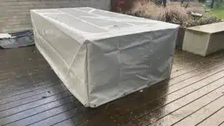 Ein rechteckiger Gartentisch mit einer beigefarbenen rechteckigen Abdeckhaube steht auf einem nassen Holzdeck. In der Nähe sorgen eine Betonbank und ein Pflanzkübel mit Gräsern für Charme, alles umgeben von einer Backsteinmauer und einem Metallzaun.