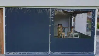 Ein Carport oder Terrassenplanke mit einer großen dunkelblauen Plane und einem klaren Fensterbereich umschließt die Terrasse teilweise und gibt den Blick auf zwei Holzstühle und einen Tisch auf einem grauen Ziegelpflaster frei.