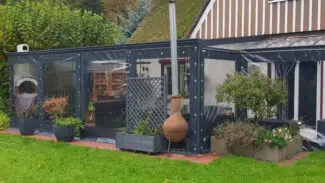 Eine gemütliche Terrasse mit transparenter Carport- oder Terrassenplane – mit 1x Fenster/ohne Tür verfügt über Hängelampen, einen Kaminofen und Topfpflanzen. Das reetgedeckte Haus liegt eingebettet zwischen Bäumen und üppigem Grün.