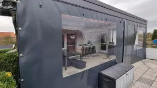 Eine überdachte Terrasse mit dunklen Planenwänden und durchsichtigen Kunststofffenstern gibt den Blick auf die Möbel im Inneren frei. Die Terrassenverkleidung, oder „Carport- oder Terrassenplane – mit 1x Fenster/ohne Tür“, befindet sich auf einem gefliesten Außenbereich unter grauem Himmel.