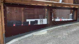 Ein Außensitzplatz mit Carport- oder Terrassenplane – mit klaren Kunststofffenstern in dunklen Rahmen unter einem Holzdach. Im Inneren sind Tische mit schwarzen Stühlen und roten Kissen zu sehen, daneben eine Leiter und eine Laterne.