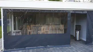 Eine transparente Carport- oder Terrassenplane umschließt die Terrasse und schützt die Barhocker und Tische aus Holz. Ein großer Heizstrahler wärmt den gefliesten Boden. Rechts vervollständigen eine weiße Wand und eine kleine Treppe die Szene.