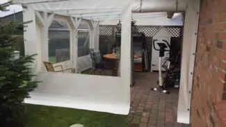 Auf der Terrasse eines Backsteingebäudes steht ein weißer Carport oder eine Terrassenplane mit Fenster und Tür. Holzstühle, ein Tisch und Trainingsgeräte stehen auf dem Backsteinboden neben einem kleinen Schuppen und einem immergrünen Baum.