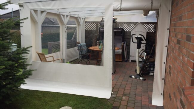 2tt (2) Auf der Terrasse eines Backsteingebäudes steht ein weißer Carport oder eine Terrassenplane mit Fenster und Tür. Holzstühle, ein Tisch und Trainingsgeräte stehen auf dem Backsteinboden neben einem kleinen Schuppen und einem immergrünen Baum.