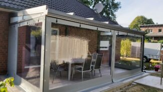 Eine Terrasse mit Carport- oder Terrassenplane – mit 1x Fenster/ohne Tür verfügt über transparente Vinylvorhänge, die einen Sitzbereich im Freien umschließen, der an ein Backsteinhaus mit grauem Dach angeschlossen ist, umgeben von Bäumen und einem in der Nähe geparkten LKW.