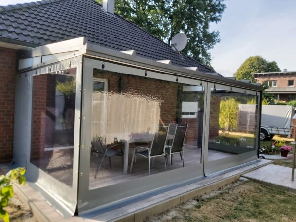 Eine Terrasse mit Carport- oder Terrassenplane – mit 1x Fenster/ohne Tür verfügt über transparente Vinylvorhänge, die einen Sitzbereich im Freien umschließen, der an ein Backsteinhaus mit grauem Dach angeschlossen ist, umgeben von Bäumen und einem in der Nähe geparkten LKW.