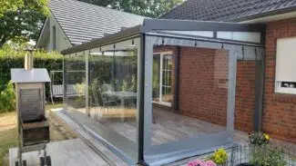 Ein Wintergartenanbau mit großen Glasflächen und einer Carport- oder Terrassenplane ist an ein Backsteinhaus angebaut. In der Nähe schmücken bunte Topfblumen den Bereich neben einem Außengrill und einem Teich, der von üppigem Grün umgeben ist.