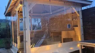 Ein hölzernes Gartenhaus mit transparenten Kunststoffplatten und einer Terrassenverkleidung - Carport- oder Terrassenplane mit 1x Fenster/ohne Tür steht in der Abenddämmerung warm beleuchtet auf einer Terrasse neben einem Tisch und einer Lampe.