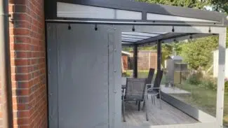 Eine überdachte Terrasse mit Carport- oder Terrassenplane und einem Fenster umschließt einen Essbereich mit Stühlen um einen Tisch mit Metallrahmen. Neben einer Backsteinmauer bietet er Ausblicke ins Grüne.
