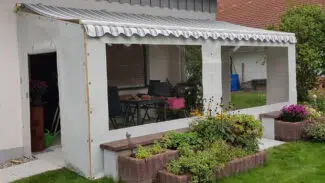 Ein Carportplane mit zwei Fenstern umschließt eine überdachte Terrasse inmitten eines üppigen Gartens. Gestreifte Markisen und klare Seiten laden ein, während blühende Pflanzen und braune Ziegelpflanzgefäße den Raum wunderschön einrahmen.