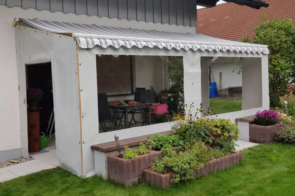 Ein Carportplane mit zwei Fenstern umschließt eine überdachte Terrasse inmitten eines üppigen Gartens. Gestreifte Markisen und klare Seiten laden ein, während blühende Pflanzen und braune Ziegelpflanzgefäße den Raum wunderschön einrahmen.