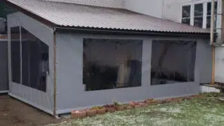 Eine graue Carport- oder Terrassenplane aus Kunststoff mit schrägem Metalldach und zwei transparenten Fenstern, die an einem Gebäude befestigt ist, steht in einem verschneiten Hof mit einem angrenzenden Blumenbeet.