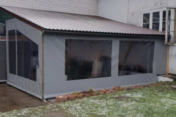 Eine graue Carport- oder Terrassenplane aus Kunststoff mit schrägem Metalldach und zwei transparenten Fenstern, die an einem Gebäude befestigt ist, steht in einem verschneiten Hof mit einem angrenzenden Blumenbeet.