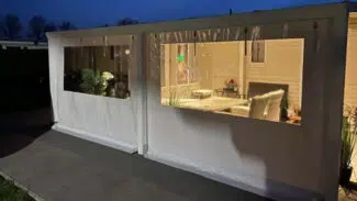 Ein gemütlicher Innenraum mit hellen Holzwänden, Korbmöbeln und Pflanzen erstrahlt bei Dämmerung durch große Fenster. Daneben bietet ein Carport mit 2-Fenster-Überdachung einen geschützten Blick auf die charmante Steinterrasse.