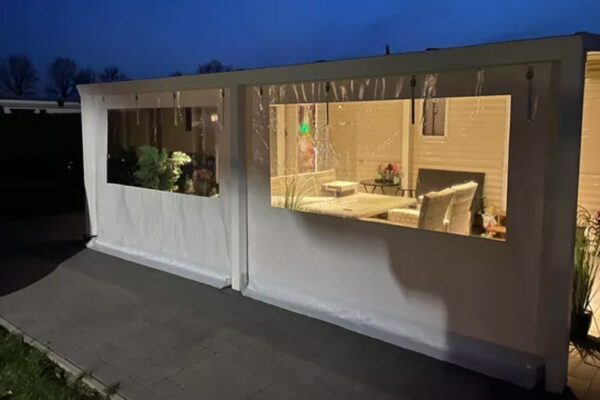 Ein gemütlicher Innenraum mit hellen Holzwänden, Korbmöbeln und Pflanzen erstrahlt bei Dämmerung durch große Fenster. Daneben bietet ein Carport mit 2-Fenster-Überdachung einen geschützten Blick auf die charmante Steinterrasse.