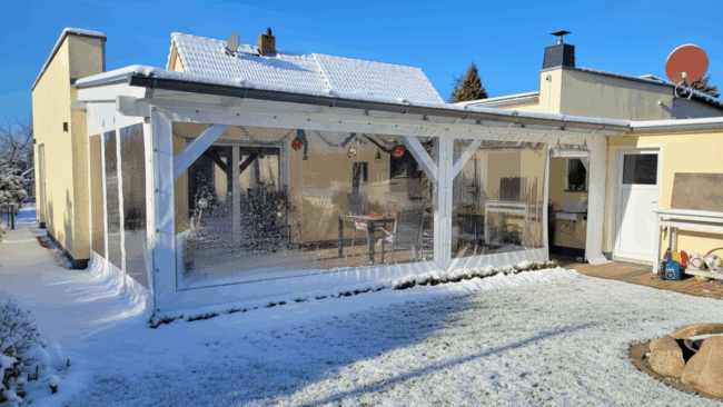 Eine überdachte Terrasse mit einem Vordach und einer Terrassenverkleidung - Carport- oder Terrassenplane mit 1x Fenster/ohne Tür, sowie ein schneebedeckter Rasen.