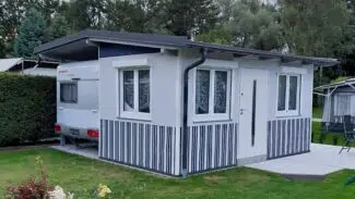 Ein kleines weiß-graues Fertighaus mit gestreiften Paneelen steht auf einem gepflegten Rasen. Daneben ist ein Wohnwagen mit der eleganten MEHGIES® VALMEX® Solera Vorzeltplane vor Bäumen unter einem bewölkten Himmel teilweise sichtbar.