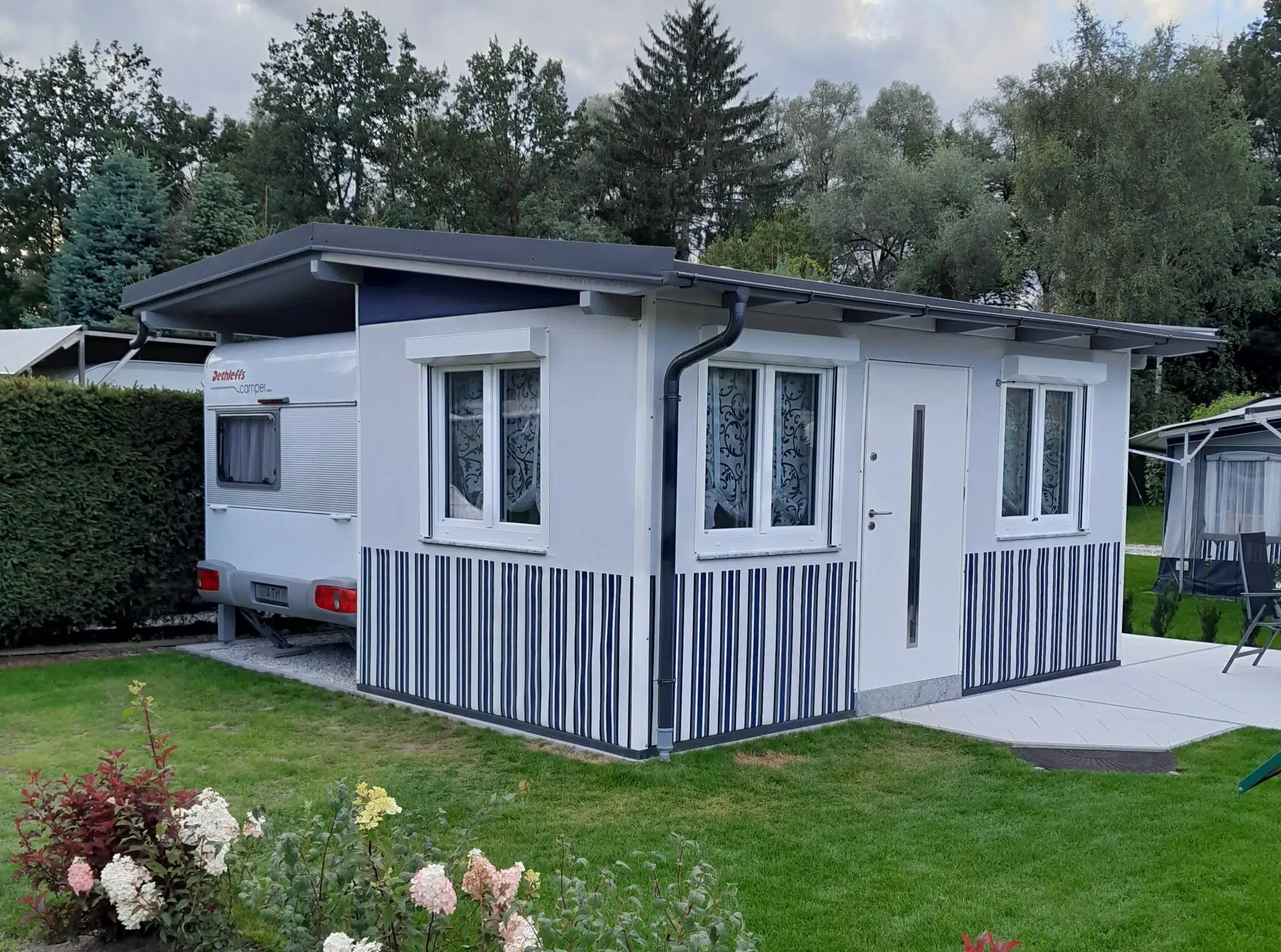 Ein kleines weiß-graues Fertighaus mit gestreiften Paneelen steht auf einem gepflegten Rasen. Daneben ist ein Wohnwagen mit der eleganten MEHGIES® VALMEX® Solera Vorzeltplane vor Bäumen unter einem bewölkten Himmel teilweise sichtbar.