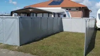 Ein Backsteinhaus mit Solarmodulen auf einem roten Dach, umgeben von einem Camping-Windschutz/Sichtschutz in einem grasbewachsenen Innenhof. Darüber ragt ein blauer Himmel mit Wolken hervor und ein gepflasterter Gehweg ziert den Vordergrund.