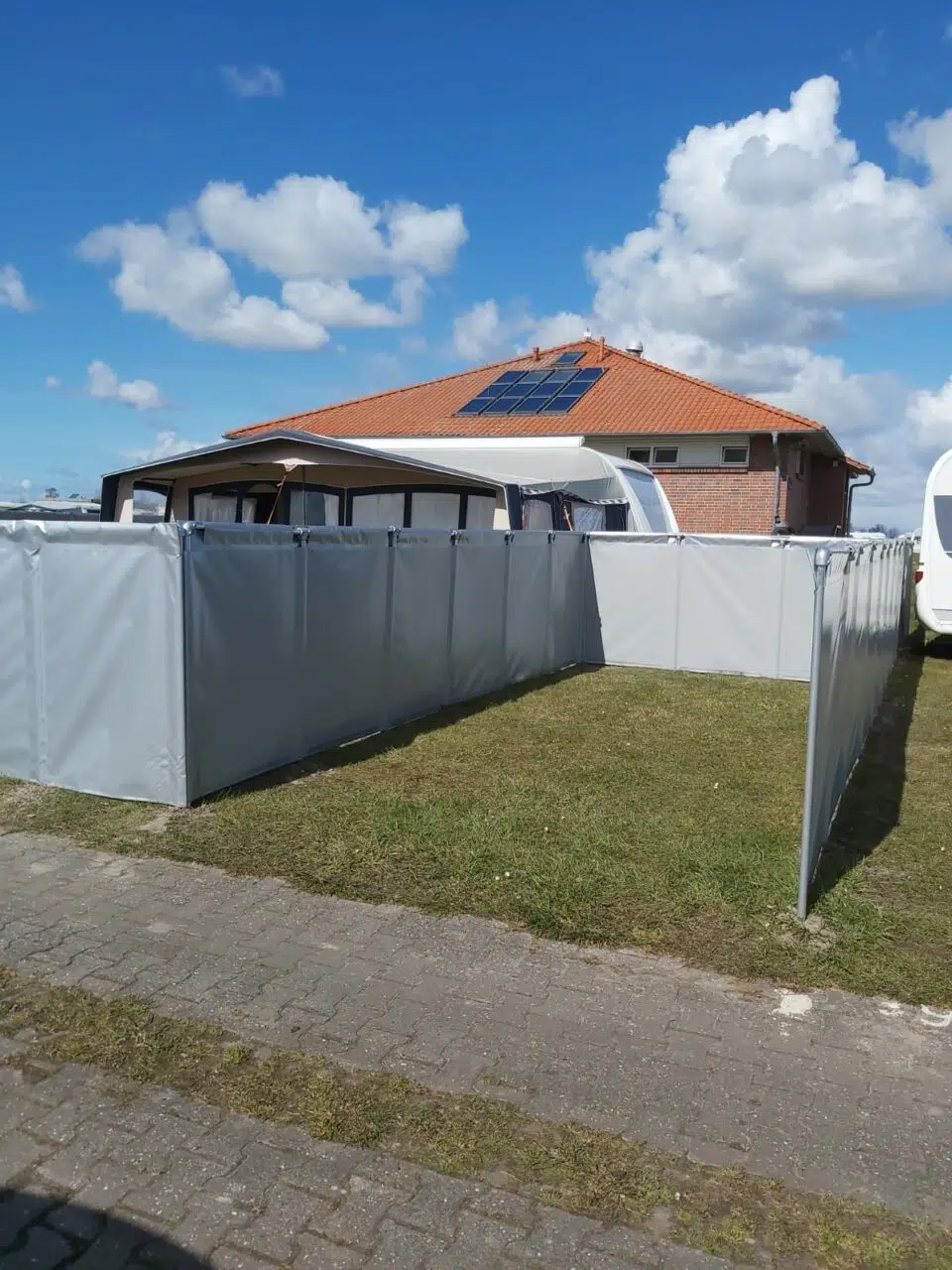 Ein Backsteinhaus mit Solarmodulen auf einem roten Dach, umgeben von einem Camping-Windschutz/Sichtschutz in einem grasbewachsenen Innenhof. Darüber ragt ein blauer Himmel mit Wolken hervor und ein gepflasterter Gehweg ziert den Vordergrund.
