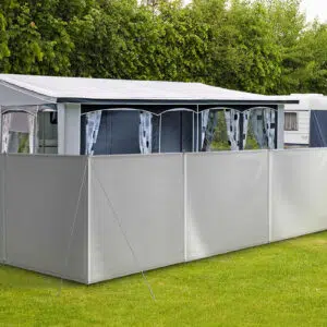 Ein Wohnwagen mit ausgefahrenem Vorzelt und grauem Camping Windschutz/Sichtschutz steht auf einer Wiese. Er ist von üppigen grünen Büschen umgeben, im Hintergrund ist ein weiterer Wohnwagen teilweise zu erkennen.