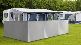 Ein Wohnwagen mit ausgefahrenem Vorzelt und grauem Camping Windschutz/Sichtschutz steht auf einer Wiese. Er ist von üppigen grünen Büschen umgeben, im Hintergrund ist ein weiterer Wohnwagen teilweise zu erkennen.