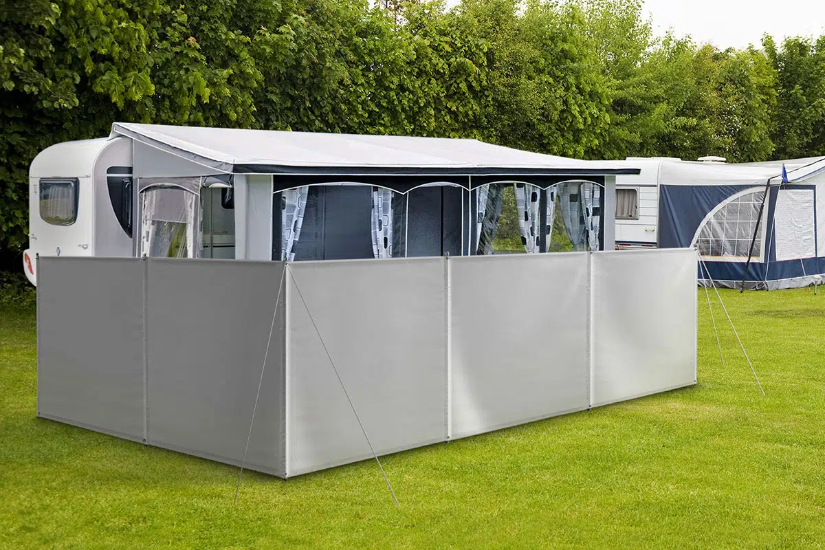 Ein Wohnwagen mit ausgefahrenem Vorzelt und grauem Camping Windschutz/Sichtschutz steht auf einer Wiese. Er ist von üppigen grünen Büschen umgeben, im Hintergrund ist ein weiterer Wohnwagen teilweise zu erkennen.