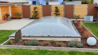 Eine Hinterhofoase verfügt über einen überdachten Pool mit rechteckigen aufblasbaren Poolplanen, umgeben von gepflegtem Gras, kleinen Bäumen und Zierpflanzen. Umgeben von einem Holzzaun sind im Hintergrund zwei Häuser zu sehen.