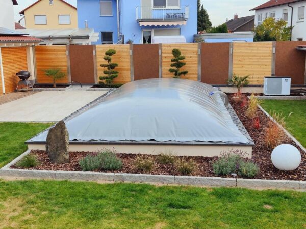 Eine Hinterhofoase verfügt über einen überdachten Pool mit rechteckigen aufblasbaren Poolplanen, umgeben von gepflegtem Gras, kleinen Bäumen und Zierpflanzen. Umgeben von einem Holzzaun sind im Hintergrund zwei Häuser zu sehen.