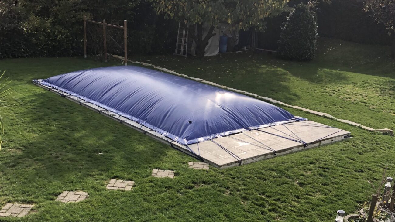 Ein rechteckiger aufblasbarer Poolplanen bedeckt einen Außenpool in einem grasbewachsenen Hof. Trittsteine führen zu dem Bereich, in dem sich Bäume und eine Leiter befinden, auf die die Sonne sanft reflektiert.