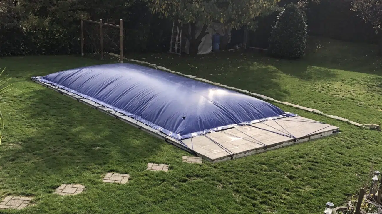 Ein rechteckiger aufblasbarer Poolplanen bedeckt einen Außenpool in einem grasbewachsenen Hof. Trittsteine führen zu dem Bereich, in dem sich Bäume und eine Leiter befinden, auf die die Sonne sanft reflektiert.