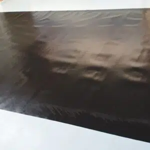 Ein großes, glänzendes schwarzes Blatt liegt flach auf einer blassen Oberfläche. Seine Textur ist glatt und leicht gewellt und reflektiert nahe Lichtquellen wie die glänzende Außenseite eines LKW-Flugzeugs in der Sonne.