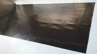 Ein großes, glänzendes schwarzes Blatt liegt flach auf einer blassen Oberfläche. Seine Textur ist glatt und leicht gewellt und reflektiert nahe Lichtquellen wie die glänzende Außenseite eines LKW-Flugzeugs in der Sonne.