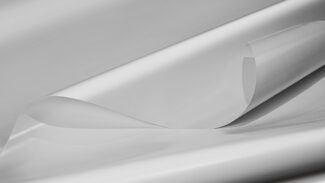 Nahaufnahme eines mehrlagigen weißen Stoffes mit glatter, glänzender Textur. Die oberste Schicht ist leicht angehoben und zeigt eine halbtransparente Qualität. Das Material wirkt leicht und zart.