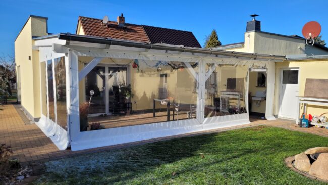 Ein Hinterhof mit einem Carport- oder Terrassenplane - mit Tür (und Fenster) auf einem weißen Rahmen, neben einem beigen Haus unter einem klaren blauen Himmel.
