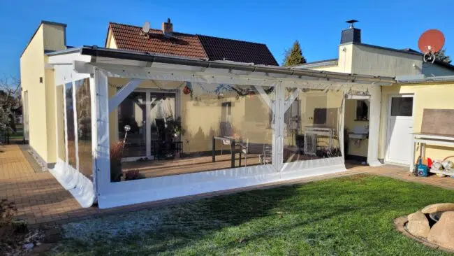 Ein Hinterhof mit einem Carport- oder Terrassenplane - mit Tür (und Fenster) auf einem weißen Rahmen, neben einem beigen Haus unter einem klaren blauen Himmel.