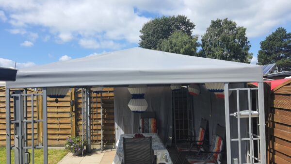 Ein Hinterhof-Setup mit Pavillondach 1 mit einem Tisch mit Tischdecke und Stühlen. Bunte Papierlaternen hängen vom Baldachin, umgeben von Holzzäunen, üppigen Bäumen und einem blauen Himmel.