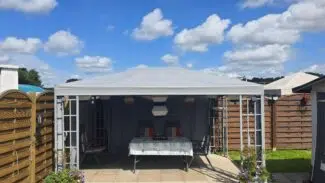 Das Pavillondach 2 bietet mit seinem PVC-Plandach unter strahlend blauem Himmel Schutz für den Terrassentisch und die Stühle. Holzzäune und Gras rahmen die Szene ein, während zwei Hängelampen das Dekor bereichern.