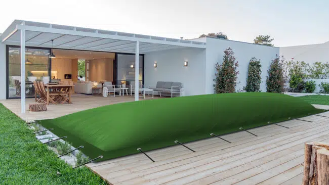 Ein moderner Hinterhof verfügt über eine überdachte Terrasse mit einer Pergola aus Holz. Zu den Gartenmöbeln gehören ein Esstisch und ein Sofa. Eine große, grüne, aufgeblasene Poolabdeckung steht auf einem Holzdeck und ist mit Gurten befestigt. Das umgebende Grün verleiht Charme.