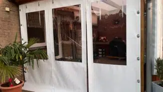 Die Carport- oder Terrassenplane mit 3 Fenstern und 1 Tür hängt an einer Metallstange und schafft einen gemütlichen Terrassenplatz zwischen Topfpflanzen und Backsteinmauern.