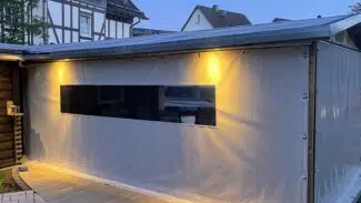 Eine überdachte Außenterrasse mit Schiebeplanenwänden, zwei gelben Lichtern und einem langen rechteckigen Fenster in der Abenddämmerung, umgeben von Häusern.