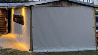 Eine hölzerne Außenstruktur mit beigen Schiebeplanen an den Seiten, beleuchtet von warmen gelben Lichtern entlang des Weges und im Inneren.