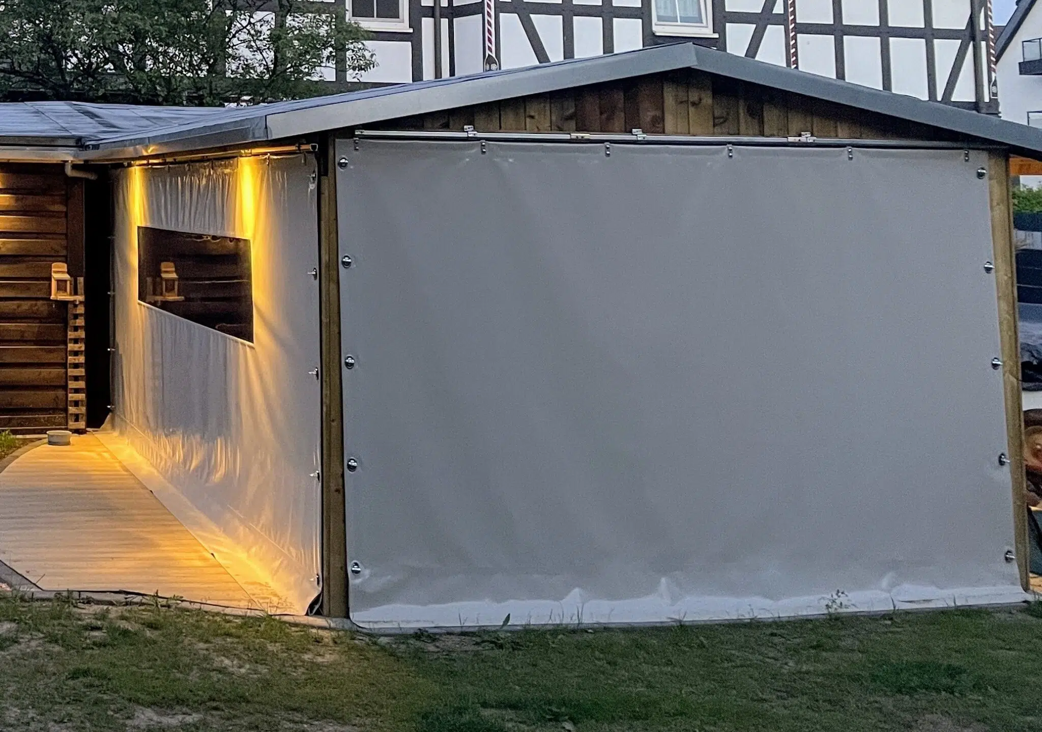 Eine hölzerne Außenstruktur mit beigen Schiebeplanen an den Seiten, beleuchtet von warmen gelben Lichtern entlang des Weges und im Inneren.